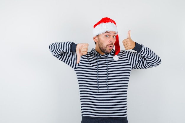 Giovane che mostra i pollici verso il basso e verso l'alto, aprendo la bocca in felpa con cappuccio, cappello da Babbo Natale e guardando stupito.