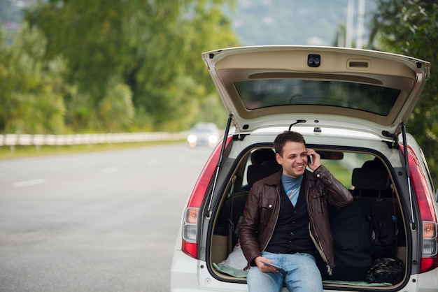 giovane che parla sul telefono cellulare e che si siede su un tronco di automobile