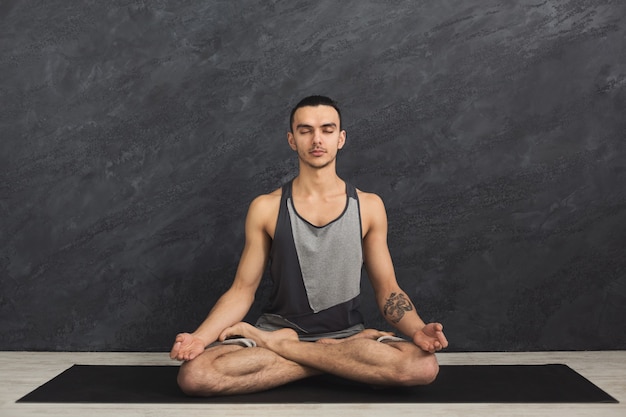 Giovane che pratica yoga, esercizi di meditazione. Il ragazzo fa la posa del loto per rilassarsi, seduto sul tappetino in palestra. Stile di vita sano, copia spazio