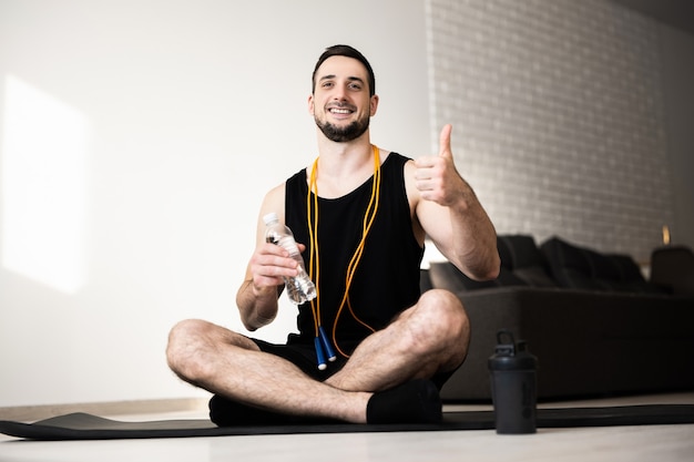 Giovane che si esercita a casa. Il ragazzo si siede da solo sulla mappa yoga nera, tiene la bottiglia d'acqua e il pollice in alto. Corda per saltare gialla intorno al collo. L'atleta riposa e si rilassa. Piccola pausa.