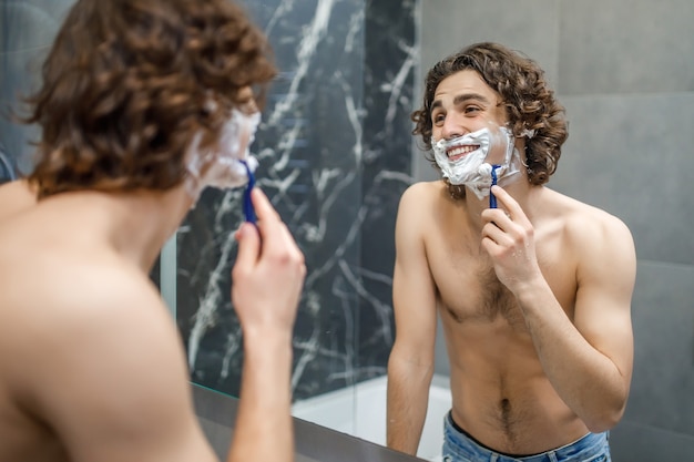 Giovane che si fa la barba vicino allo specchio nel bagno moderno