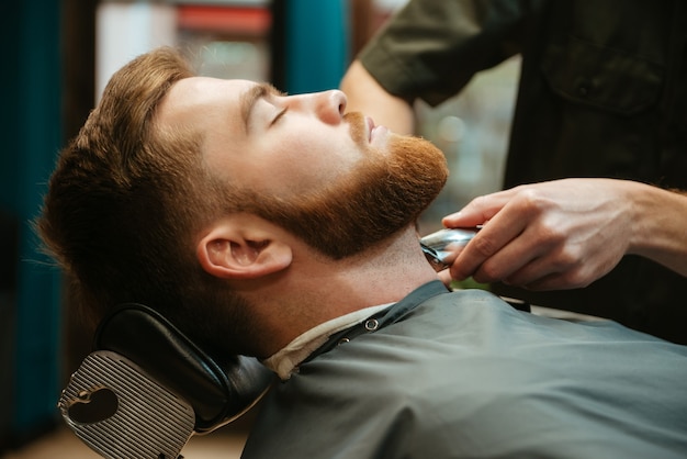 giovane che si fa tagliare la barba dal parrucchiere mentre si trova sulla sedia al barbiere.