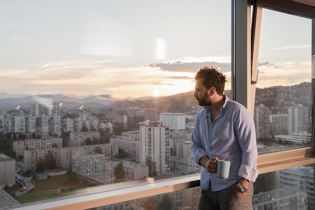 giovane che si gode il caffè serale e il bellissimo paesaggio al tramonto della città mentre si trova vicino alla finestra