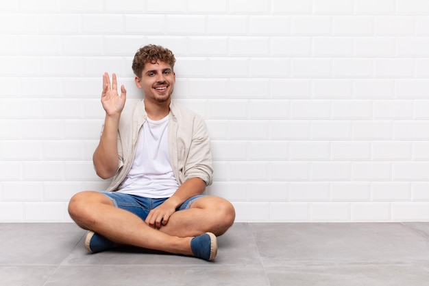 Giovane che sorride allegramente e allegramente, agitando la mano, dandoti il benvenuto e salutandoti o salutandoti