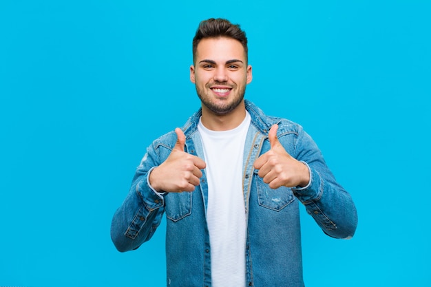 Giovane che sorride con gioia e che sembra felice, sentendosi spensierato e positivo con entrambi i pollici su contro il fondo blu