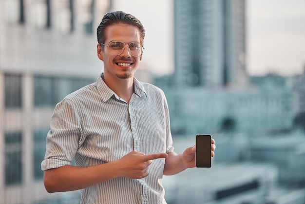 Giovane che tiene in mano un telefono, felice con un sorriso sul viso, che mostra il dito sul telefono