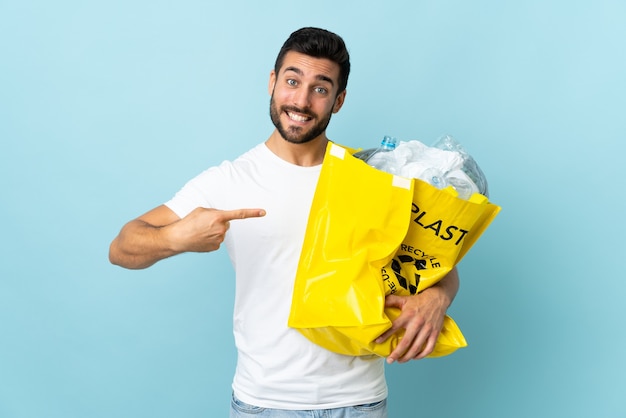 Giovane che tiene un sacchetto pieno di bottiglie di plastica da riciclare isolato sulla parete blu e indicandolo