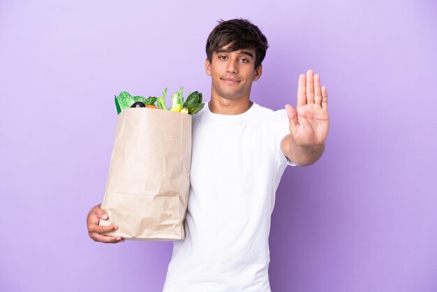 Giovane che tiene una borsa della spesa isolata su sfondo viola che fa un gesto di arresto