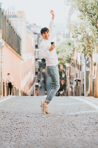 Giovane che tiene una tazza di caffè che celebra e balla in strada. Concetti di libertà e tecnologia, stile moderno a madrid