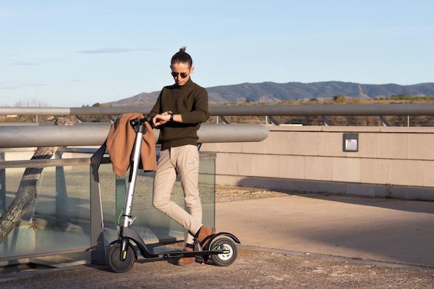 Giovane che usa lo smartwatch e si muove con uno scooter elettrico nel parco cittadino con le montagne dietro e lo spazio della copia