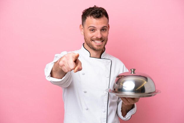 Giovane chef brasiliano con vassoio isolato su sfondo rosa che punta davanti con felice espressione