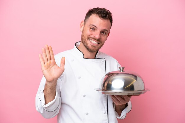 Giovane chef brasiliano con vassoio isolato su sfondo rosa che saluta con la mano con espressione felice