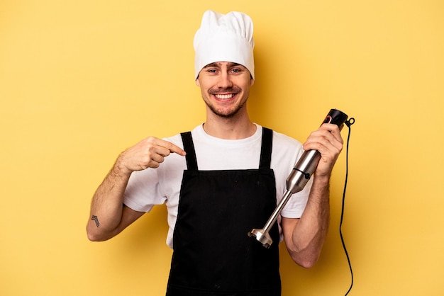 Giovane chef caucasico che tiene il frullatore isolato su sfondo giallo persona che indica a mano uno spazio per la copia della camicia, orgoglioso e fiducioso