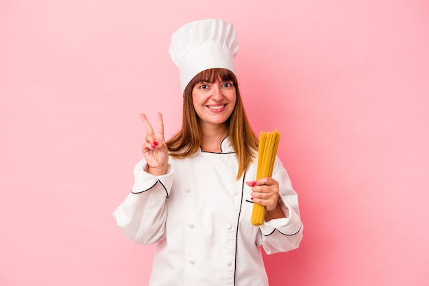 Giovane chef caucasico donna che tiene la pasta isolata su sfondo rosa che mostra il numero due con le dita.