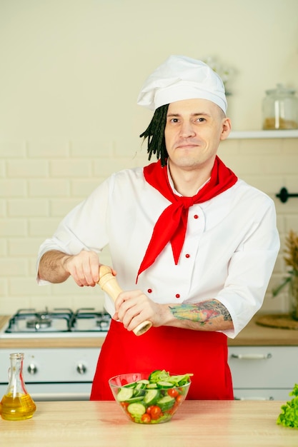 Giovane chef con dreadlocks infarcendo insalata di verdure in cucina Uomo sorridente in uniforme preparare insalata macinando spezie con mulino