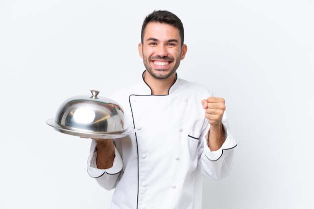 Giovane chef con vassoio isolato su sfondo bianco che celebra una vittoria nella posizione del vincitore