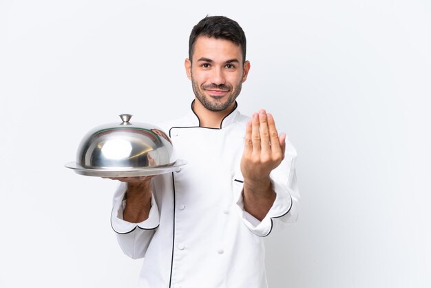 Giovane chef con vassoio isolato su sfondo bianco che invita a venire con la mano Felice che tu sia venuto