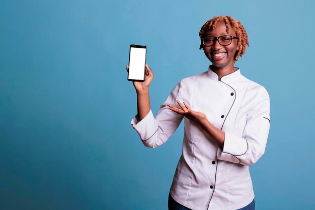 Giovane chef di cucina dai capelli ricci con gli occhiali, che mostra felicemente le ricette sullo schermo dello smartphone. Giovane donna afroamericana, fornello professionale che tiene telefono cellulare portatile.