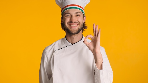 Giovane chef di successo vestito in uniforme che mostra bene il segno alla fotocamera su sfondo giallo Attraente chefcook ha approvato il piatto per la cena