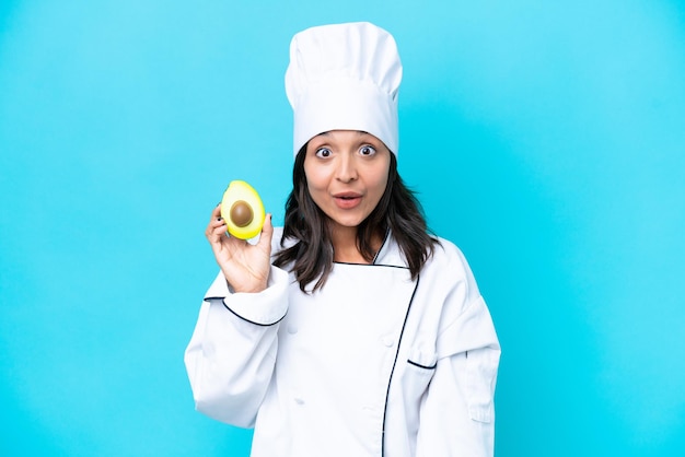 Giovane chef ispanico donna che tiene avocado isolato su sfondo blu con espressione facciale a sorpresa