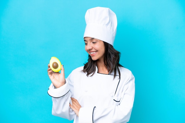 Giovane chef ispanico donna che tiene avocado isolato su sfondo blu guardando di lato e sorridente