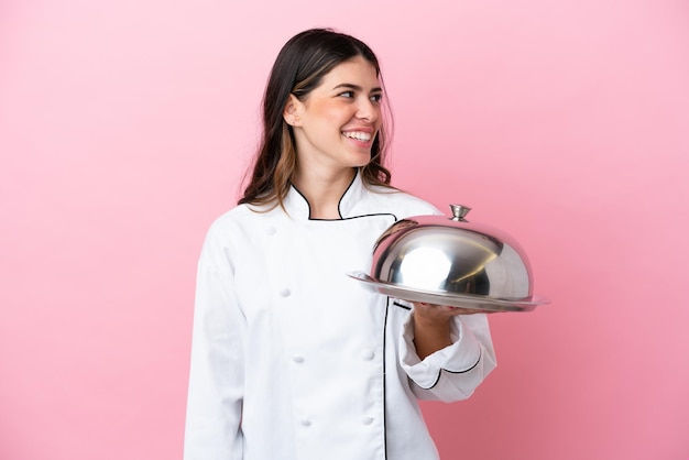 Giovane chef italiano donna che tiene vassoio con coperchio isolato su sfondo rosa guardando il lato