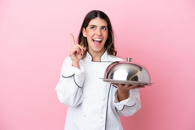 Giovane chef italiano donna che tiene vassoio con coperchio isolato su sfondo rosa pensando a un'idea che punta il dito verso l'alto