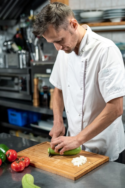 Giovane chef maschio in uniforme che si china sul tavolo mentre taglia zucchine verdi fresche su una tavola di legno circondata da pomodori e avocado