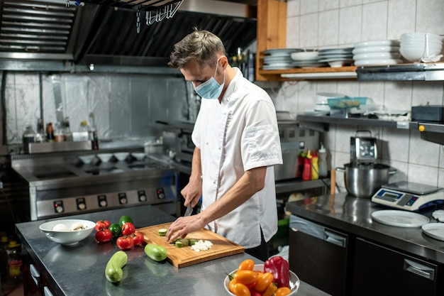 Giovane chef professionista in uniforme e maschera protettiva che si china sul tavolo in cucina mentre taglia zucchine fresche e altre verdure