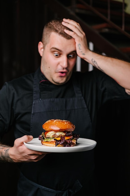 Giovane chef sorpreso che tiene un piatto con hamburger delizioso