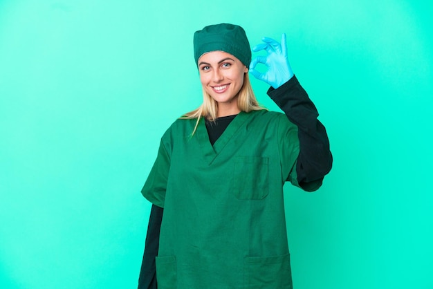 Giovane chirurgo donna uruguaiana in uniforme verde isolata su sfondo blu che mostra il segno giusto con le dita