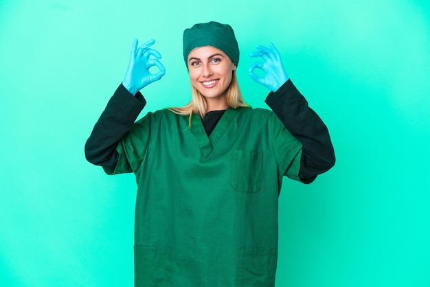 Giovane chirurgo donna uruguaiana in uniforme verde isolata su sfondo blu che mostra un segno ok con le dita