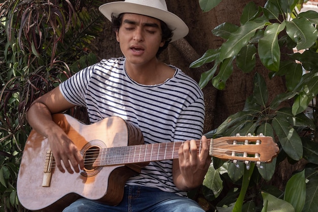 Giovane chitarrista ispanico con stile bohémien che suona la sua chitarra acustica all'aperto in un giardino del parco e canta