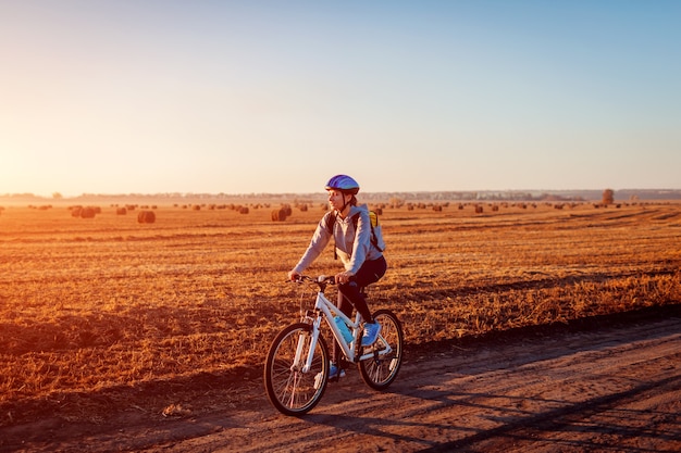 Giovane ciclista a cavallo nel campo autunnale con mucchi di fieno al tramonto. Donna con zaino in viaggio in bici. Uno stile di vita sano.