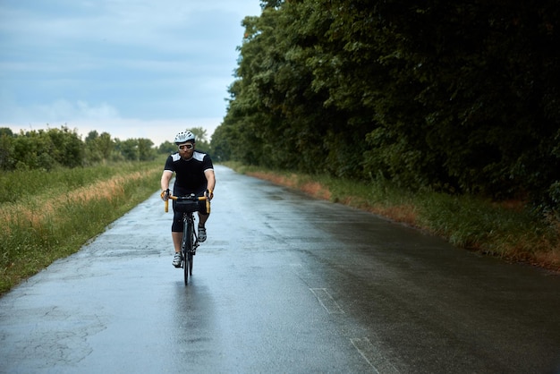 Giovane ciclista barbuto sportivo con casco e occhiali in uniforme che va in bicicletta sulla strada nel freddo