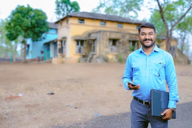 Giovane commerciante di proprietà indiano in piedi davanti alla casa con il computer portatile che parla sul telefono cellulare