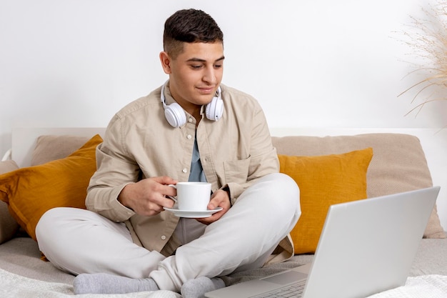 Giovane con abiti casual seduto sul letto con un computer portatile e bere tè o caffè Uomo che usa il notebook per navigare in Internet leggere notizie guardare film chiamare amici Rilassarsi a casa