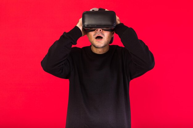 Giovane con basba corto, che indossa felpa nera, berretto e occhiali per realtà virtuale stupiti, su sfondo rosso. Concetto di tecnologia, VR, informatica e hobby.