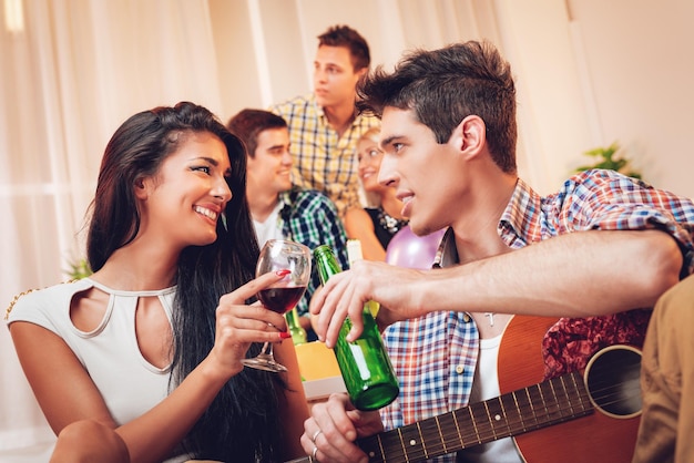 Giovane con chitarra acustica e ragazza felice che brindano con bevande a casa festa.