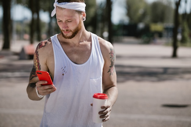 Giovane con fascia bianca vestito con una t-shirt bianca utilizzando il telefono cellulare e tiene in mano un bicchiere di plastica sul campo sportivo all'esterno in una giornata di sole.