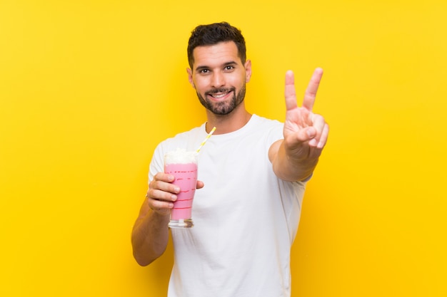 Giovane con il frappé della fragola sopra la parete gialla isolata che sorride e che mostra il segno di vittoria