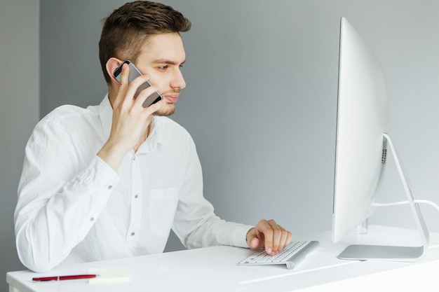 Giovane con il telefono in camicia bianca che si siede al desktop con un computer in ufficio. Lavoratore su sfondo grigio