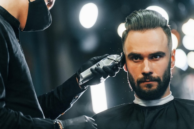 Giovane con l'acconciatura alla moda seduto e farsi rasare la barba nel negozio di barbiere