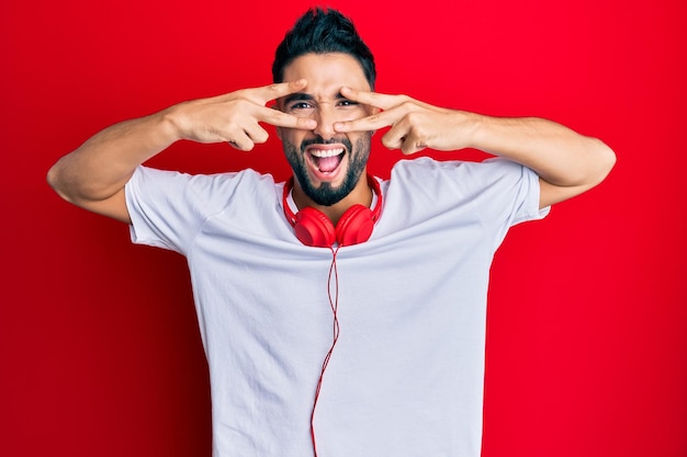 Giovane con la barba che ascolta musica utilizzando le cuffie facendo il simbolo della pace con le dita sul viso sorridente allegro mostrando vittoria