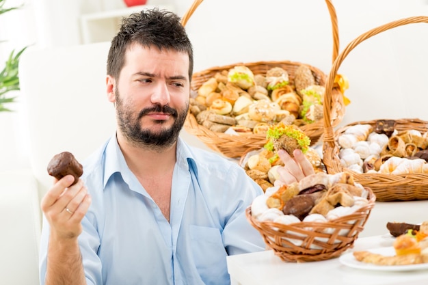 Giovane con la barba, che tiene tra le mani una pasticceria dolce e salata in cui guardare, chiedendosi chi scegliere. Sullo sfondo si possono vedere cesti intrecciati con pasticcini.