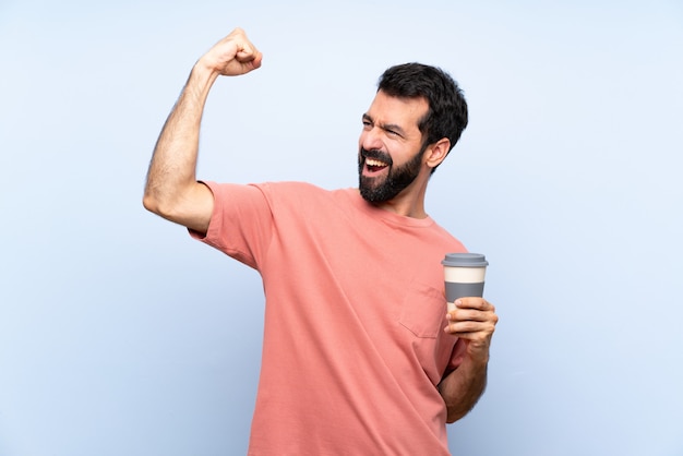 Giovane con la barba che tiene un caffè da asporto sopra il blu isolato che celebra una vittoria