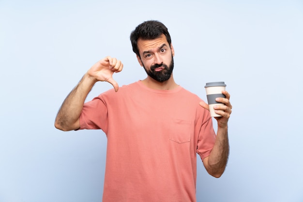 Giovane con la barba che tiene un caffè da asporto sopra il pollice di mostra blu isolato giù