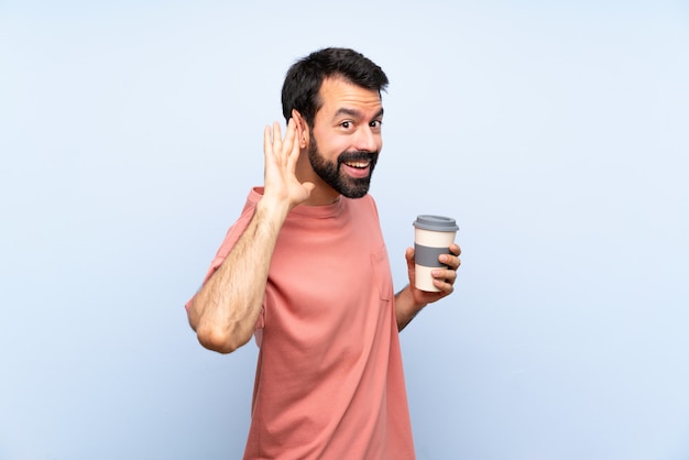 Giovane con la barba che tiene un caffè da asporto sopra la parete blu isolata che ascolta qualcosa mettendo la mano sull'orecchio