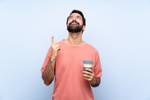 Giovane con la barba che tiene un caffè da asporto sopra la parete blu isolata che indica su e sorpreso