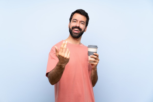 Giovane con la barba che tiene un caffè da asporto sopra la parete blu isolata che invita a venire con la mano